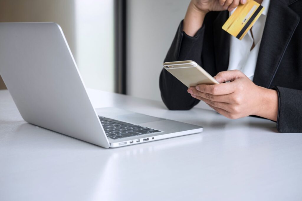 Midsection woman holding credit card while using mobile phone online shopping by laptop table