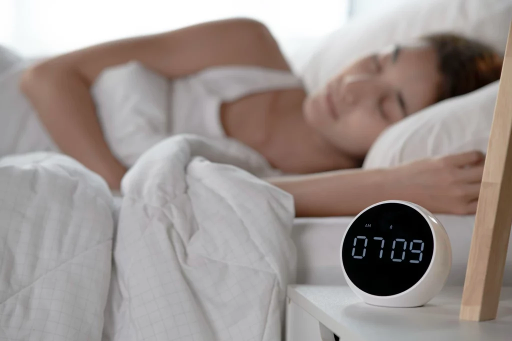 夜間臥室中，一名女性正在床上熟睡，床邊桌上放置圓形電子助眠裝置，顯示清晨時間。