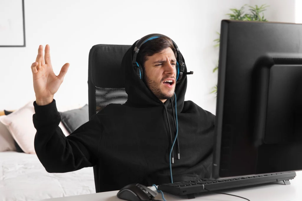 一名戴著耳機的男性玩家坐在電腦前，雙手舉起神情激動，疑似在進行線上對戰遊戲時情緒失控。